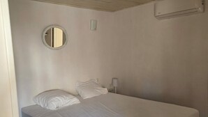 A single-bed room with a wooden ceiling, a round mirror, a bedside lamp, and a wall-mounted air conditioning unit. - Charming home in the heart of the Corbières region – Patio, courtyard. (Fabrezan)
