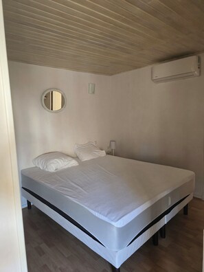 A single-bed room with a wooden ceiling, a round mirror, a bedside lamp, and a wall-mounted air conditioning unit. - Charming home in the heart of the Corbières region – Patio, courtyard. (Fabrezan)