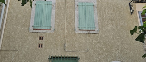 Fachada de edificio con contraventanas verdes, el número '6' visible en la puerta y plantas en macetas en la base.