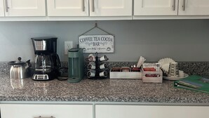 Uma bancada de cozinha com cafeteira, dispensador de água, suporte para canecas e um letreiro indicando um bar de café, chá e chocolate.