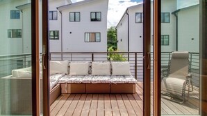 Un balcón con piso de madera, una zona de estar con cojines y una silla de metal.