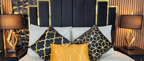 Una cama con cabecera de diseño geométrico, varios almohadones con motivos geométricos y dos lámparas de noche.