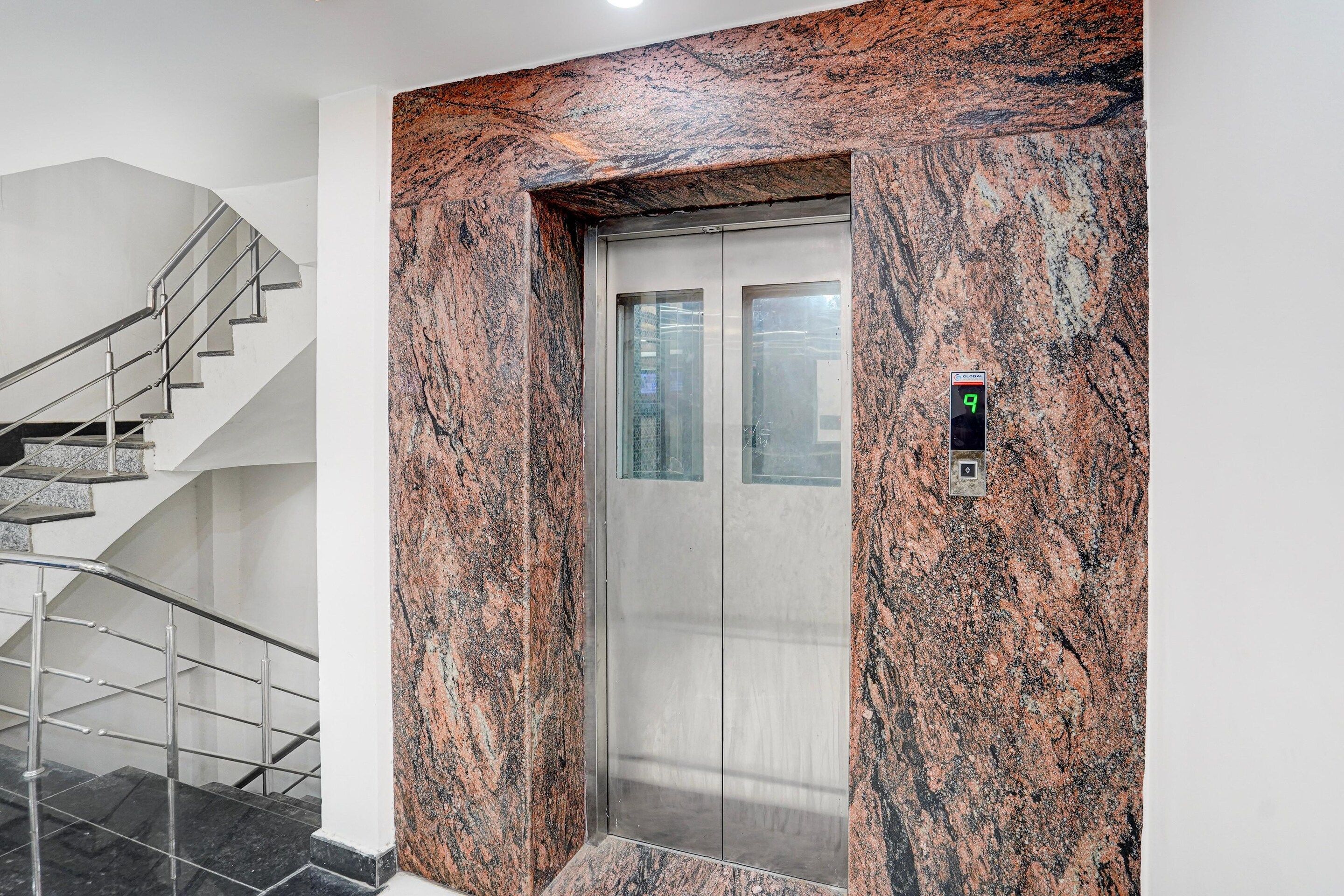 Un ascenseur avec un mur en granit, un écran numérique et un escalier avec des rampes en métal.