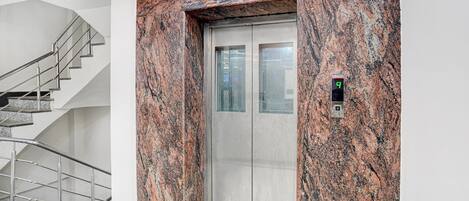 An elevator with a granite wall, a digital display, and a stairwell with metal railings.