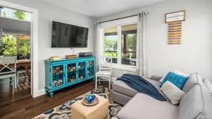 Ein Wohnzimmer mit einer Couch, einem Fernseher, einem Schrank und einem Fenster mit Blick auf Grünflächen.