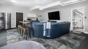 A modern living room with a sectional sofa, a flat-screen TV, a kitchen area, and a staircase.