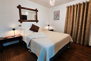 A bedroom with a bed, two bedside tables, a nightstand, a mirror, a wall-mounted light fixture, a framed picture, and a curtain.