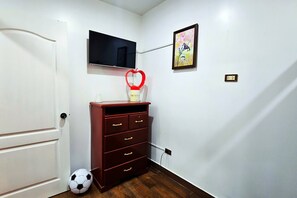 Una habitación con una puerta blanca, un televisor montado en la pared, un objeto con forma de corazón rojo sobre un tocador de madera, una pelota de fútbol en el suelo y un enchufe en la pared.