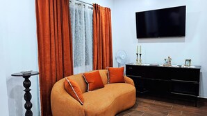 A living room with a sofa, curtains, a flat-screen TV, a fan, and a small table with decorative items.