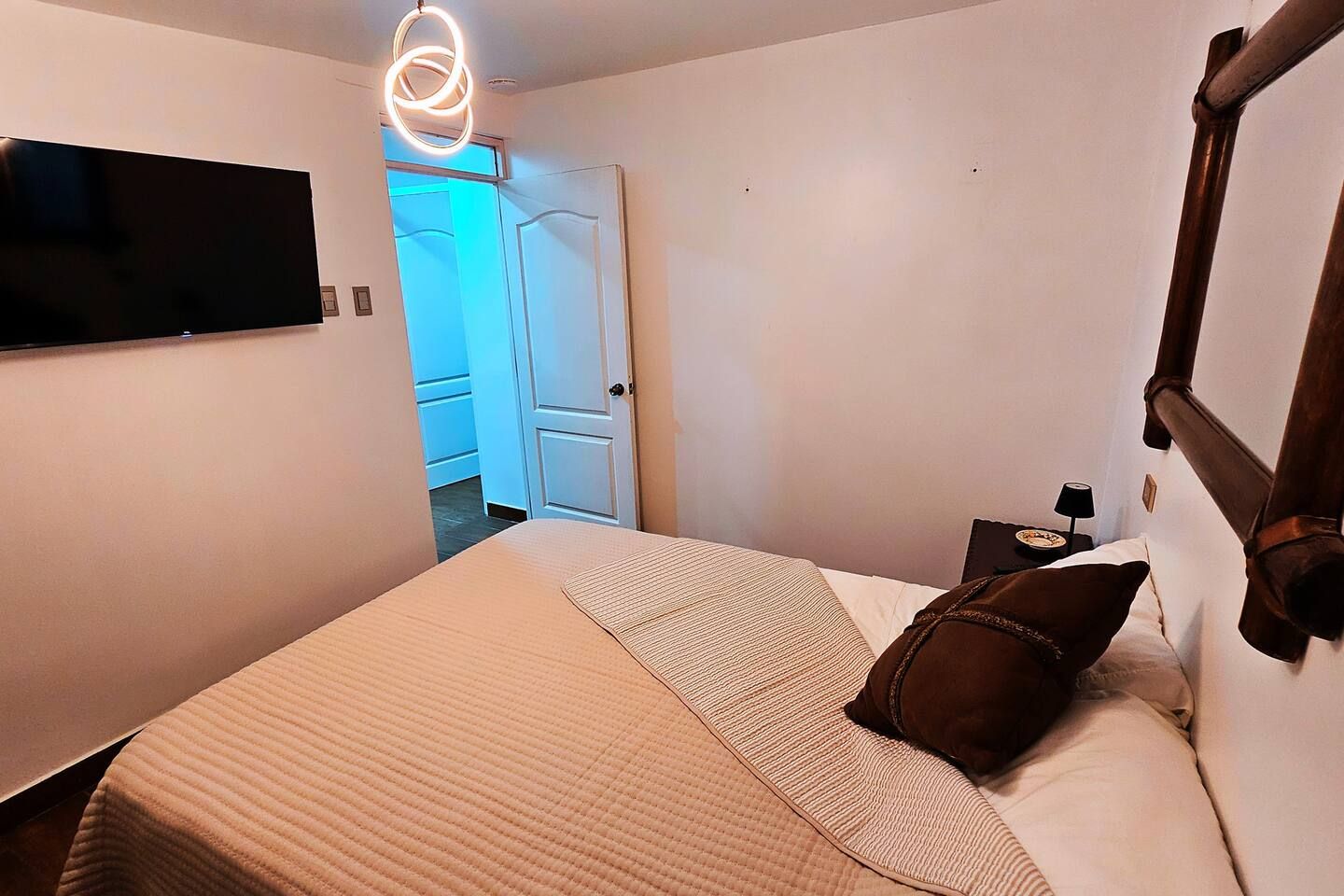 Una habitación de hotel con una cama, un televisor montado en la pared, una mesita de noche con lámpara y una puerta que da al baño.
