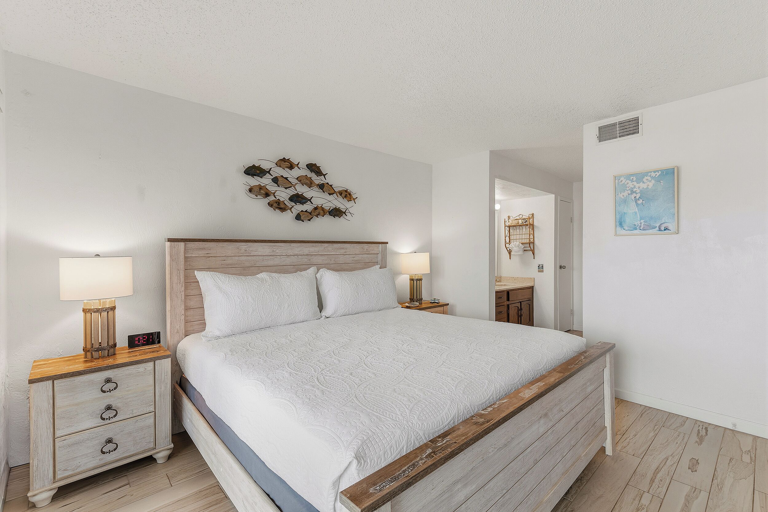 Une chambre à coucher avec une tête de lit en bois, un lit avec une literie blanche, deux tables de chevet avec des lampes, une horloge murale et un tableau décoratif représentant des poissons.