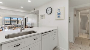 Une cuisine avec un plan de travail blanc, un évier et des placards. Il y a une horloge au mur et un tableau encadré juste au-dessus.