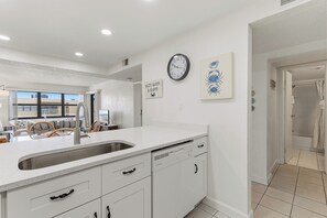 A kitchen with a white countertop, a sink, and cabinets. There is a clock on the wall and a framed picture above it. - 🌊Top Floor 1BR Condo | Ocean + Pool Views | Sleeps 4 | Castle Reef 407🌊 (New Smyrna Beach)