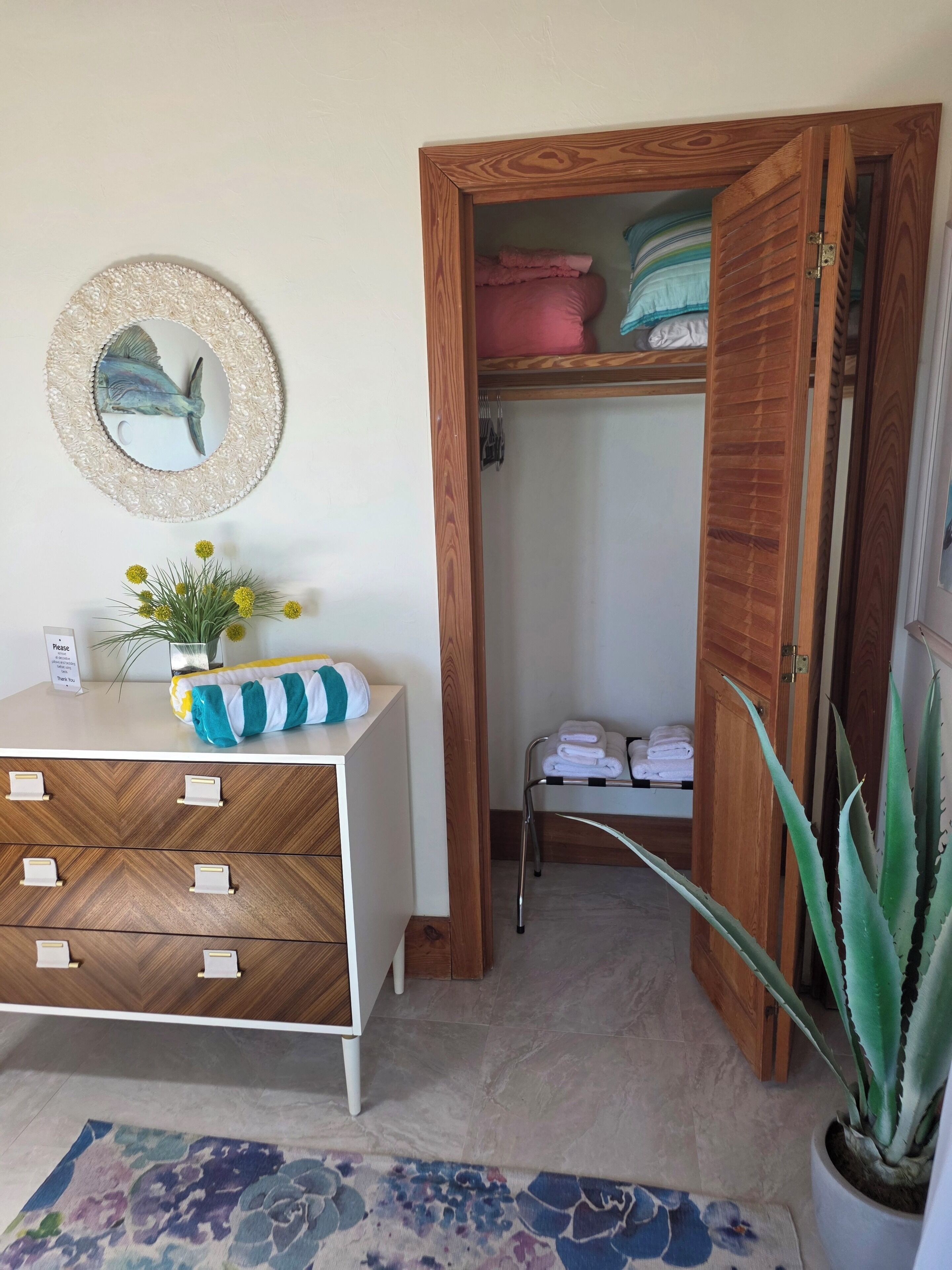 A hotel room with a wooden wardrobe, a mirror with a decorative frame, a small plant, and a floral rug.