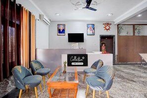 A modern living room with a glass coffee table, two blue armchairs with gold leaf patterns, a flat-screen TV mounted on the wall, and a reception desk with a sign. - Collection O Dafi Crossway Varanasi (Varanasi)