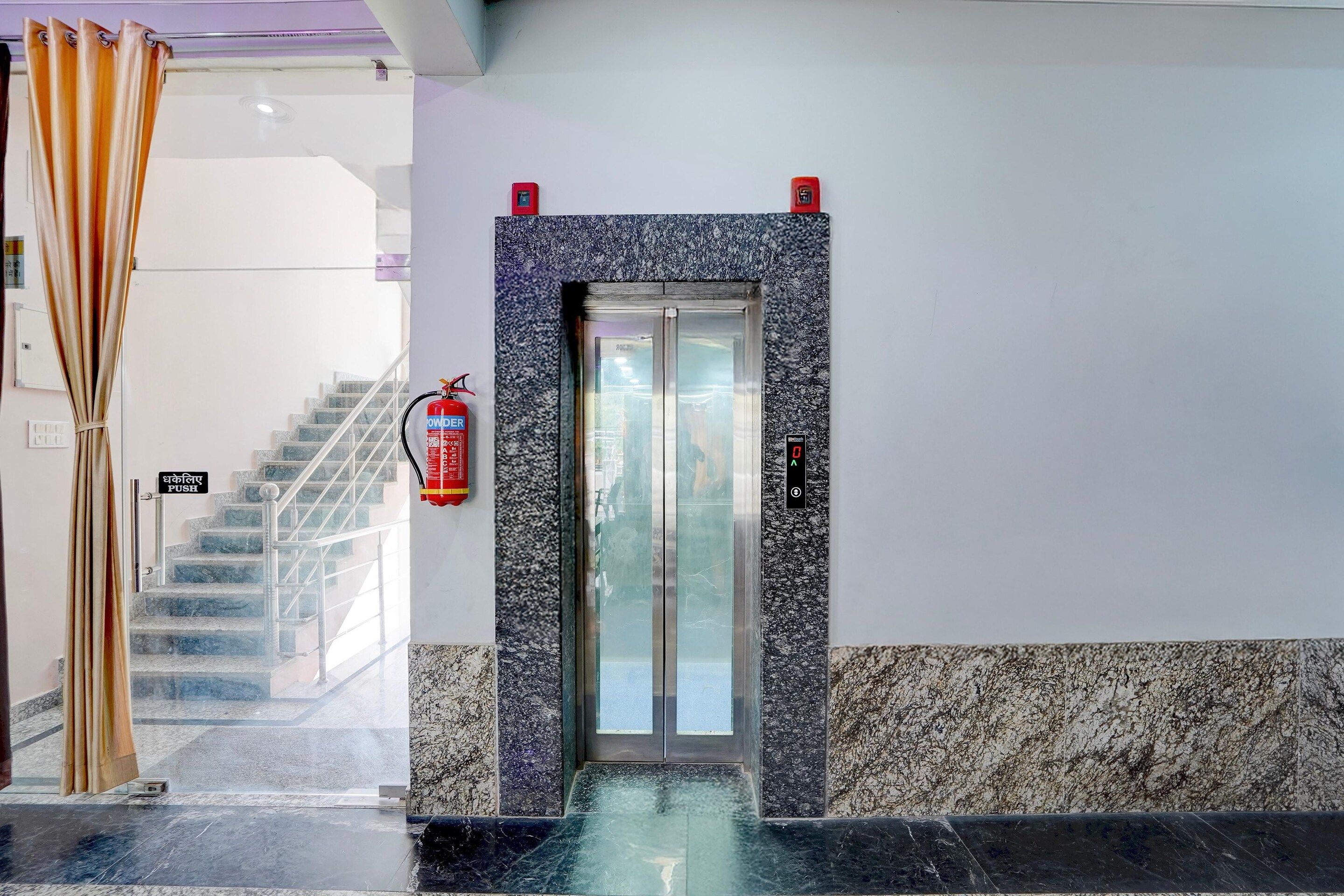 Ingresso di un hotel con parete in marmo, porta di ascensore in vetro e estintore a parete.