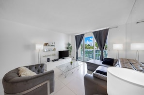 A modern living room with a sofa, a coffee table, a flat-screen TV, and a balcony with a view of palm trees and buildings. - Beautiful apartment in community with pool and beach in HOLLYWOOD BEACH MIAMI (Hollywood)