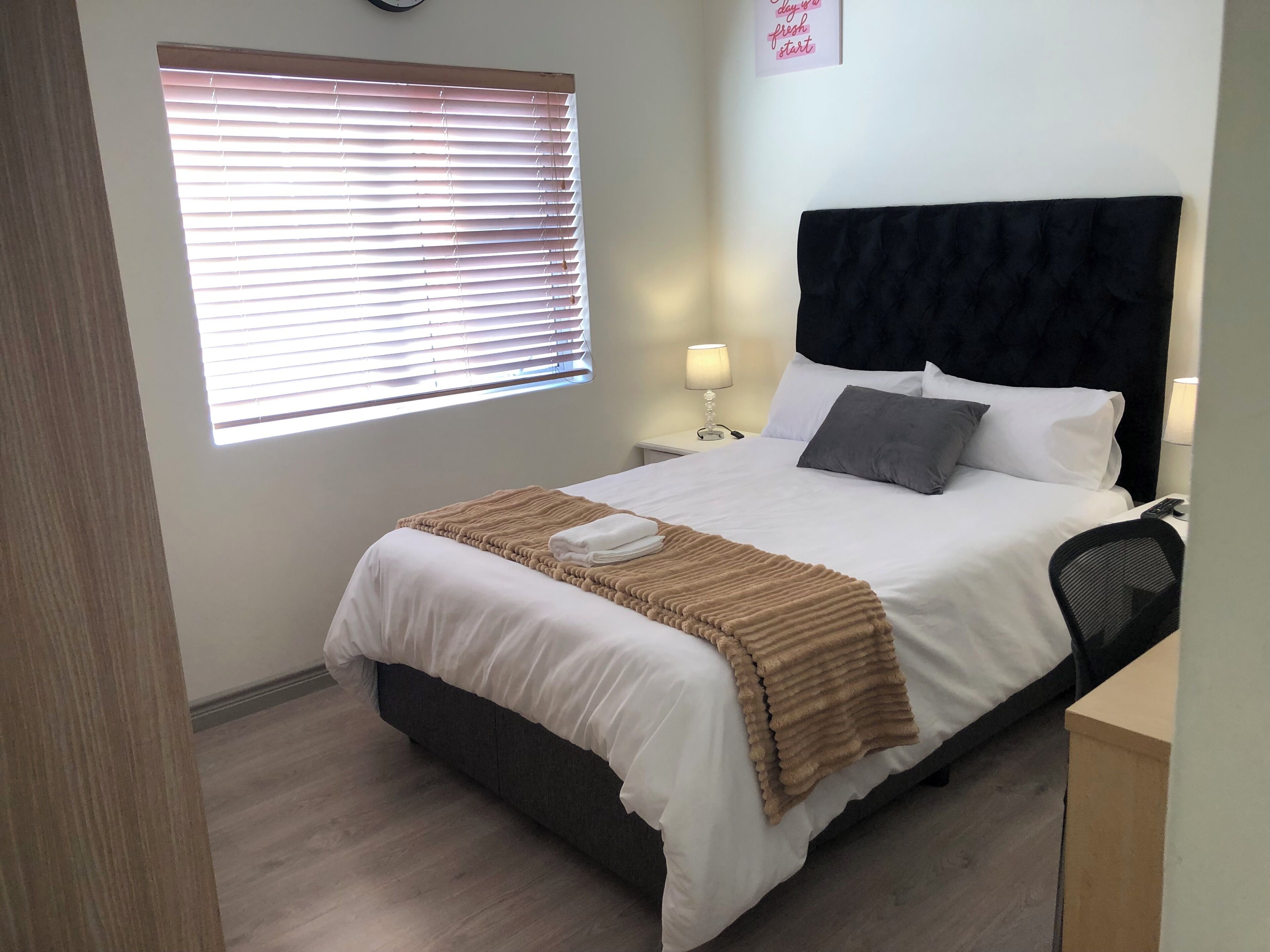 A hotel room with a bed, a nightstand, a lamp, a window with blinds, and a wall clock.