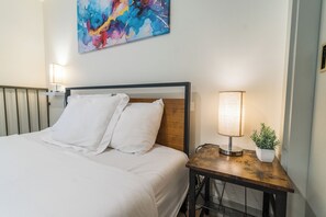 Un lit bien fait, avec du linge de lit blanc, une tête de lit en bois et une table de chevet sur laquelle se trouvent une lampe et une plante en pot.