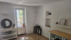 A room with a white console table, a wooden floor, a window with a view, a small table with a chair, and shelves with decorative items. - A Provençal stone farmhouse dating from 1870, with a terrace and a pool. (Mougins)