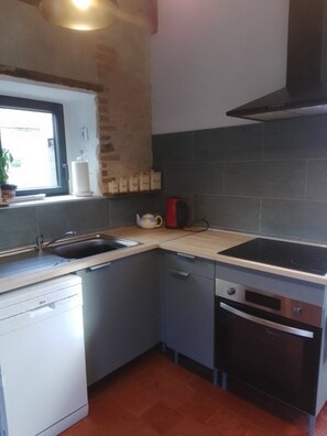 A compact kitchen with a sink, stove, and oven. There is a window with a view outside, and a shelf with kitchen items. - Charming Riverside Gite in the Brenne National Park close to amenities. (Belabre)