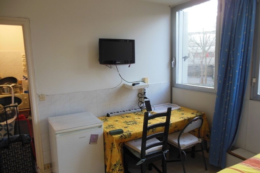 Een kleine kamer met een bed, een bureau met stoel, een aan de muur bevestigde televisie en een raam met gordijnen.