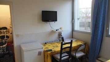 Een kleine kamer met een bed, een bureau met stoel, een aan de muur bevestigde televisie en een raam met gordijnen.