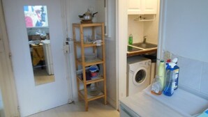 Een compacte keuken met een wasmachine, een houten plank met servies en een klein aanrecht met schoonmaakmiddelen.