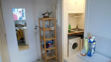 Une cuisine compacte équipée d’un lave-linge, d’une étagère en bois où sont rangés la vaisselle, et d’un petit plan de travail avec des produits de nettoyage.