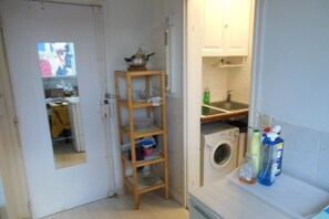 A compact kitchen with a washing machine, a wooden shelf with dishes, and a small countertop with cleaning supplies. - STUDIO APARTMENT IN CENTRAL PARIS (Paris)