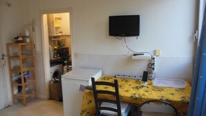 Een kleine keuken met een tafel, stoelen, een koelkast, een aan de muur bevestigde televisie en een houten plank.