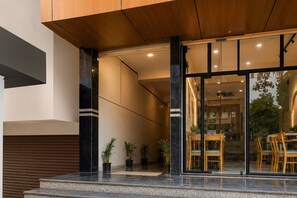 A modern building entrance with a glass door, potted plants, and a seating area visible through the glass. - Deluxe Lavish Stays (Hyderabad)