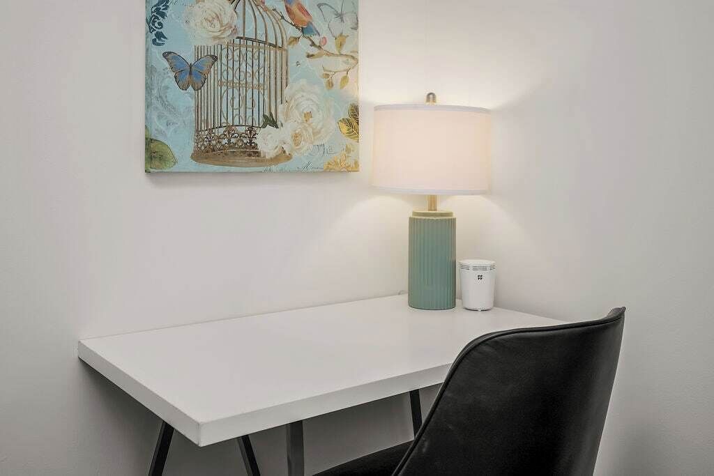 Ett vitt bord med en turkos lampa och en vit kopp bredvid, en svart stol framför, och en vägg med en fågelbur och blommotiv.