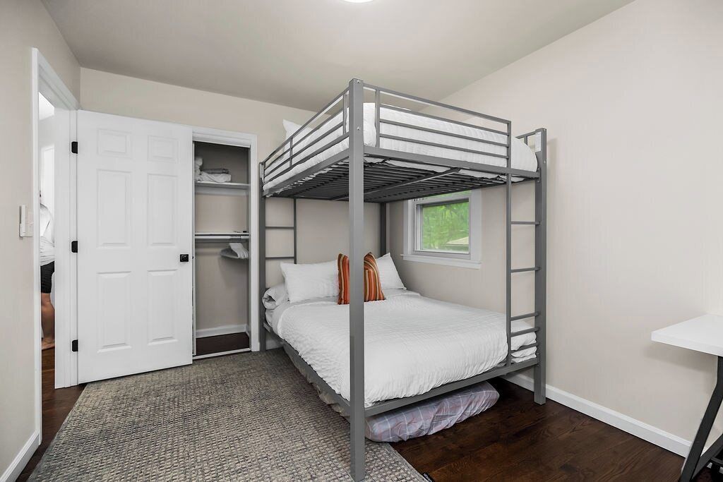 Una cama litera con escalera, una puerta blanca, un ventanal pequeño y un escritorio blanco.