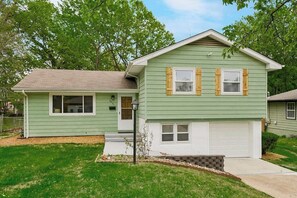 A two-story house with a front garage, a small porch, and a well-maintained lawn. - Modern 3BR, Sleeps 9, Mins to Arrowhead & KC (Raytown)