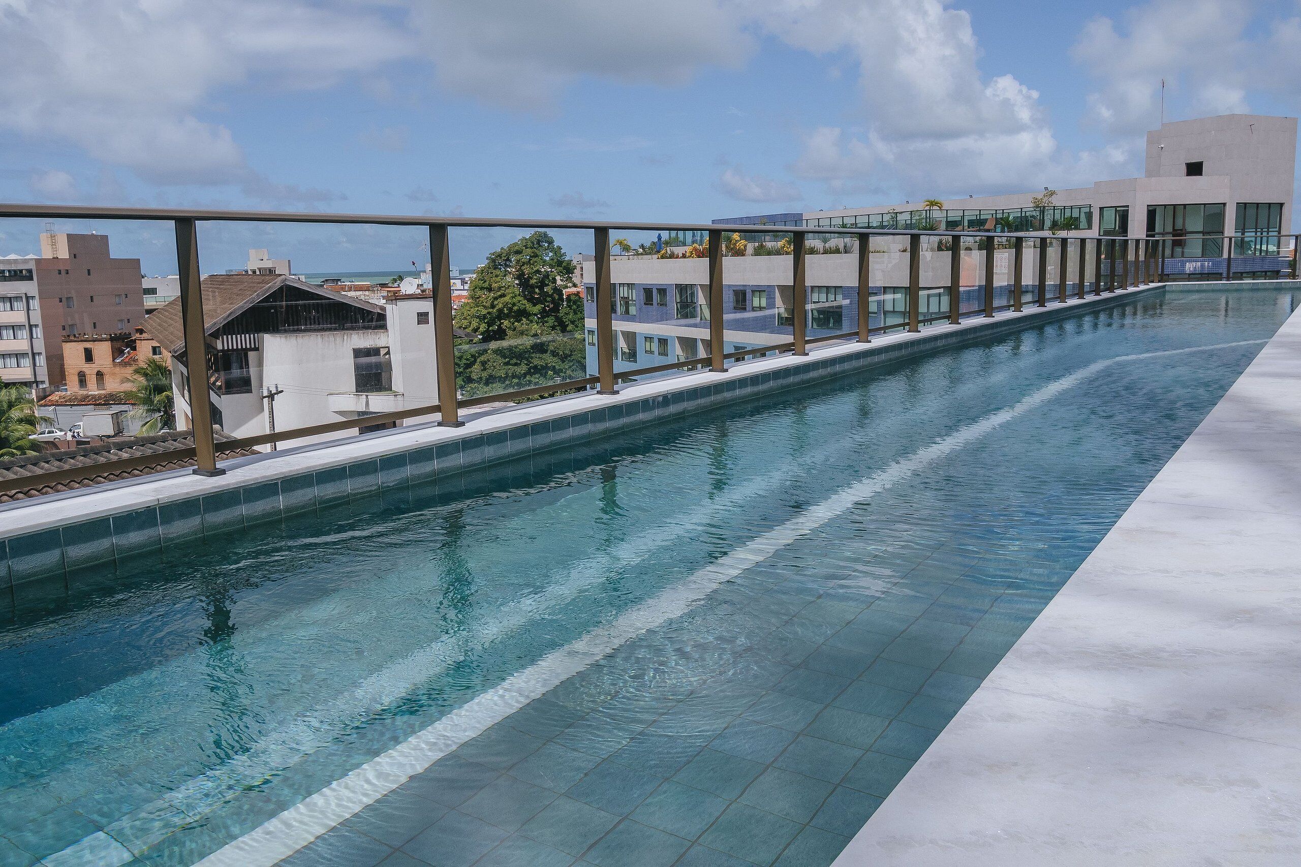 A long, narrow swimming pool with a clear blue water surface, surrounded by a modern building with large windows and a metal railing.
