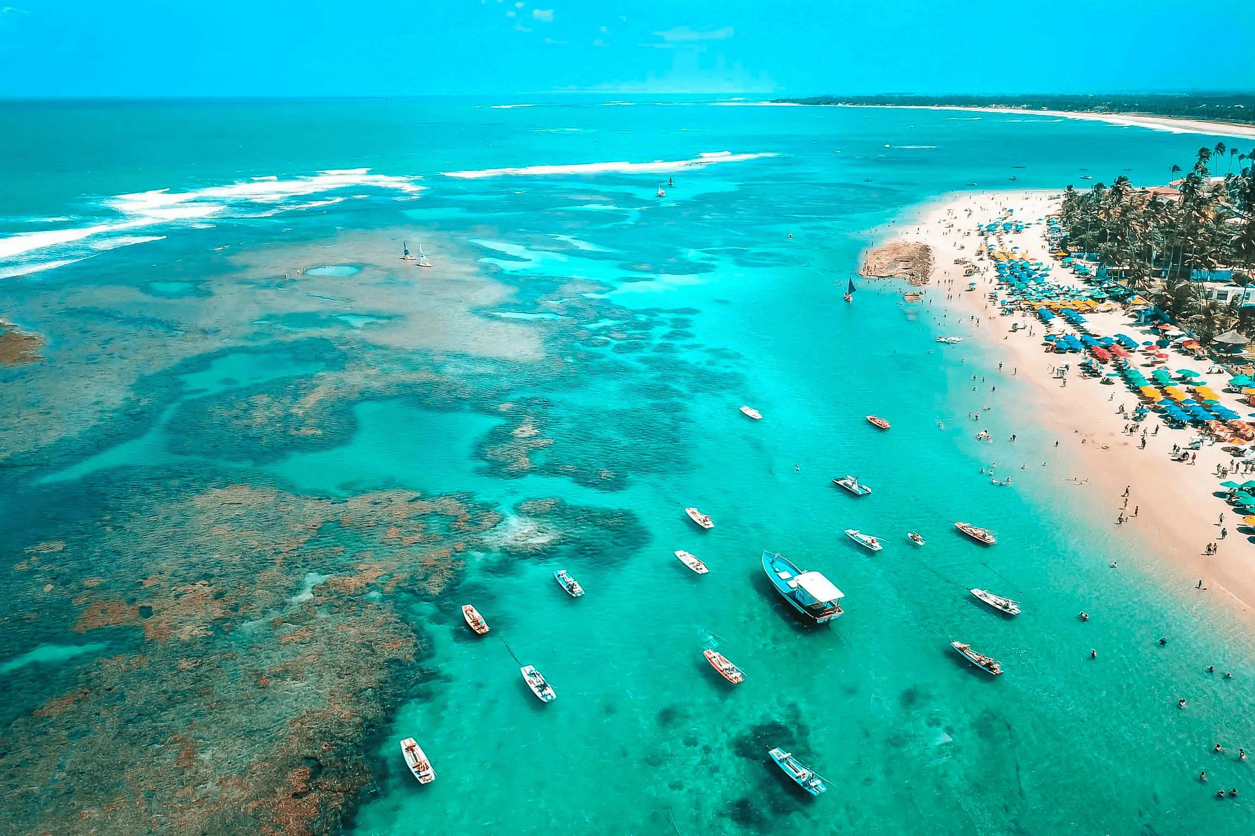 An aerial view of a beach with clear turquoise waters, boats, and a sandy shore with umbrellas.