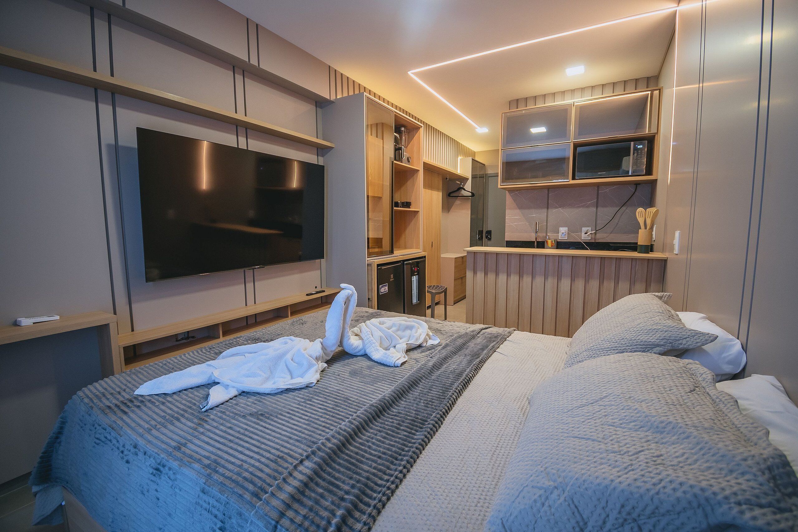 A modern hotel room with a large bed, a flat-screen TV mounted on the wall, a built-in wardrobe, and a kitchenette area.