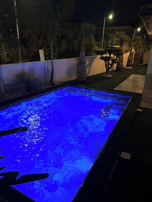 Ein rechteckiger Swimmingpool mit blauem Licht, umgeben von einem angelegten Garten mit verschiedenen Pflanzen und einem Gebäude im Hintergrund.