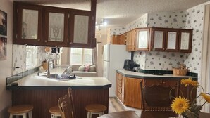A kitchen with wooden cabinets, a central island with stools, a refrigerator, and a dining area with a chair and a vase of sunflowers. - Cozy Lakehouse Gem In the Woods (Hephzibah)