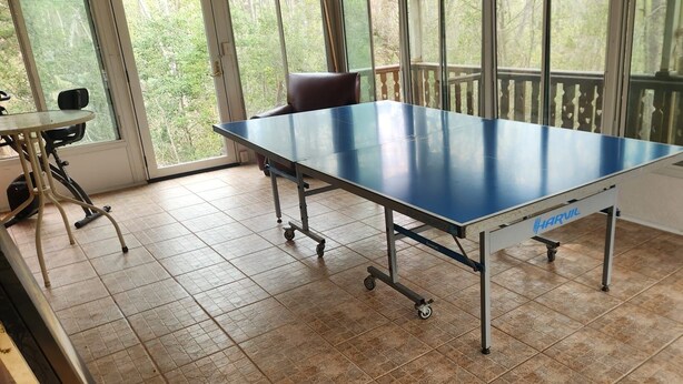 A blue table tennis table with wheels, a chair, and a small table with a chair, all in a room with large windows and a tiled floor. - Cozy Lakehouse Gem In the Woods (Hephzibah)