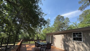 A wooden deck with a table and chairs, surrounded by trees, leading to a house with a window. - Cozy Lakehouse Gem In the Woods (Hephzibah)