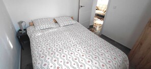 Ein kleiner, schmaler Raum mit einem Bett, einem Nachttisch und einer Tür zu einem anderen Raum.