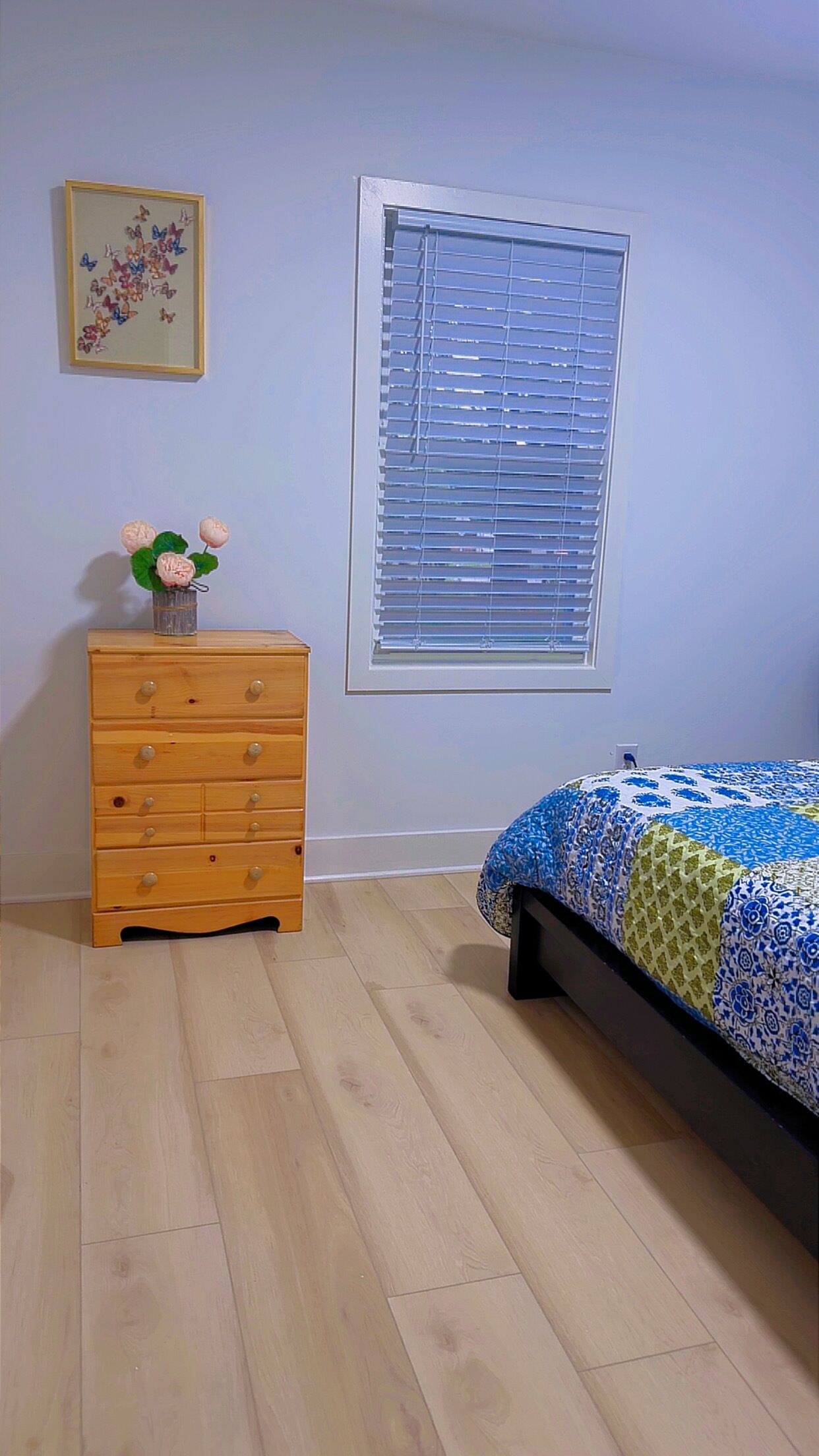 Een slaapkamer met een houten dressoir, een bed met een bedsprei met dessin, een raam met zonwering en een aan de muur hangend schilderij.