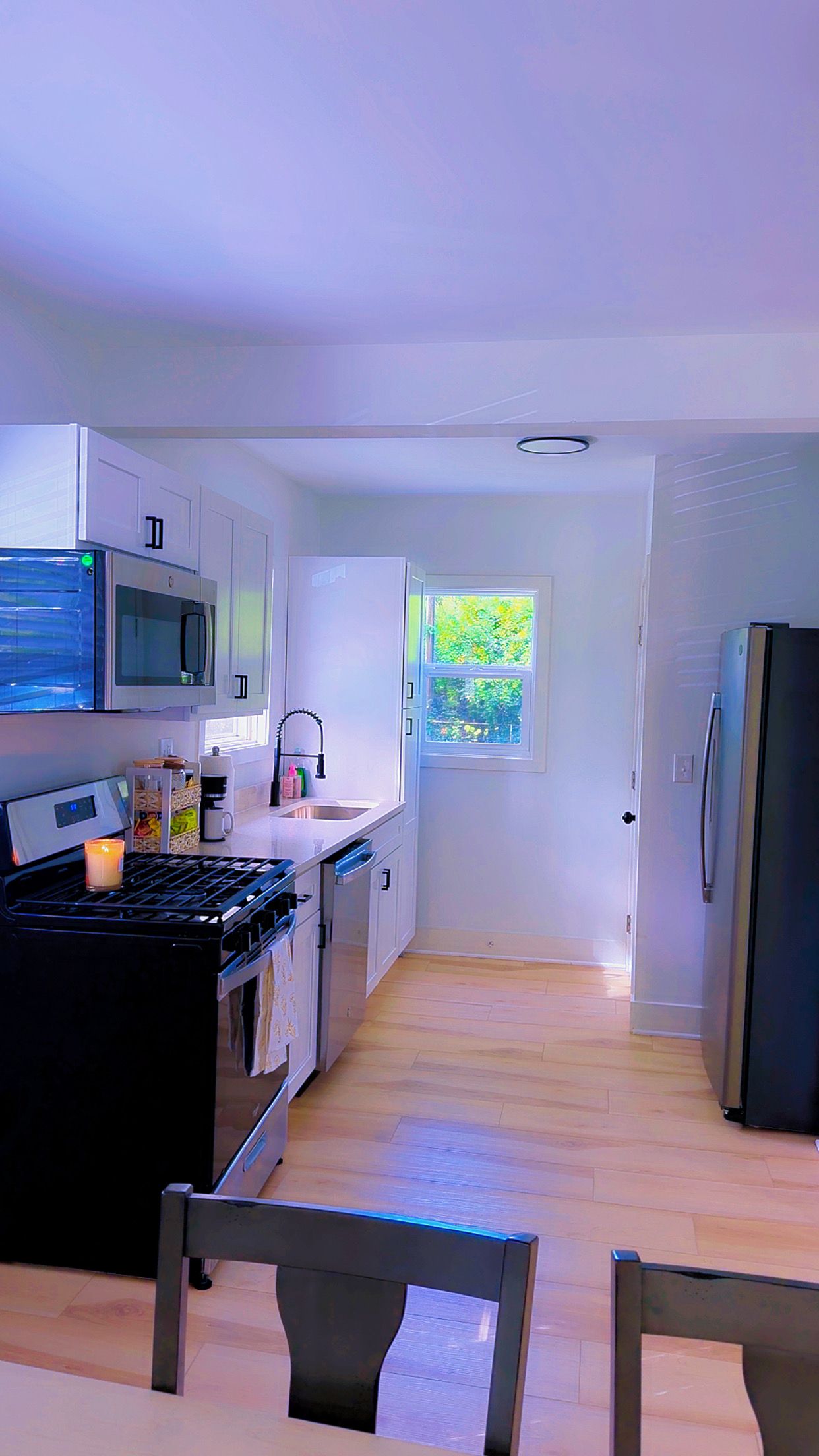 Une cuisine moderne avec des meubles blancs, une cuisinière, un four à micro-ondes et un réfrigérateur. Une fenêtre offre une vue sur la verdure environnante.