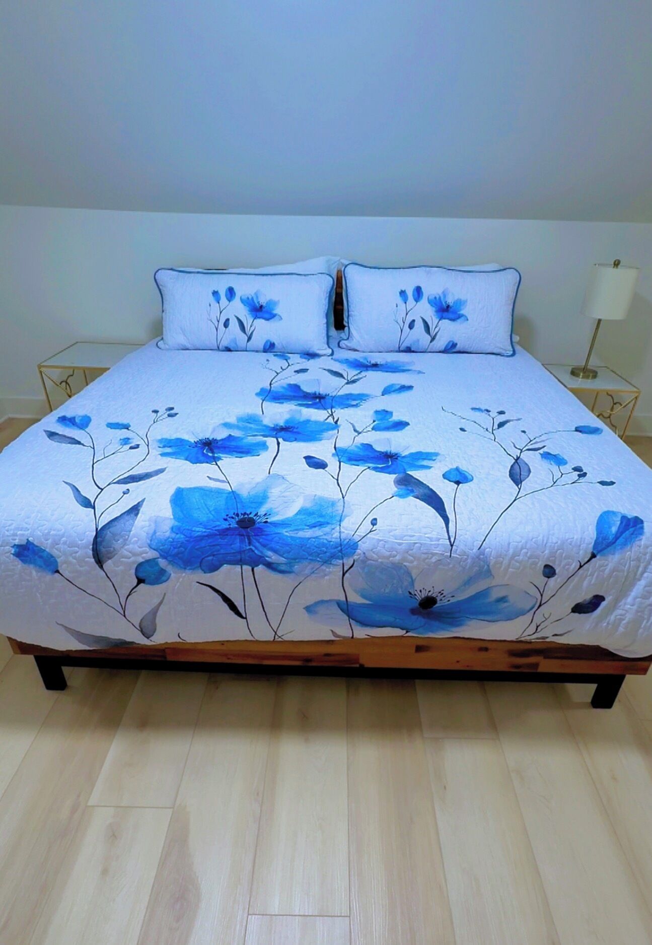 A bed with a floral bedspread and matching pillows, a bedside table with a lamp, and a wooden floor.
