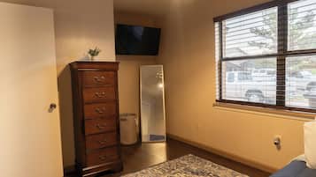 Una habitación con un aparador de madera, un televisor blanco montado en la pared, una alfombra estampada y una ventana con persianas.