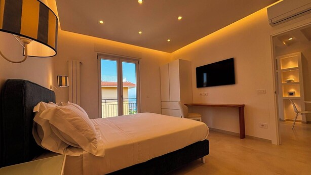 A modern bedroom with a bed, bedside table, TV, and a balcony view. - De Vivo Realty - Panorama Suite Sorrento (Sorrento)