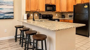 A kitchen with wooden cabinets, a granite countertop, a black refrigerator, and three bar stools. - Myrtle Beach Golf Resort Condo - Pools & Hot Tub (Myrtle Beach)
