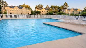 En swimmingpool med ett vitt poolområde och solstolar, omgiven av ett vitt staket och bostadshus.